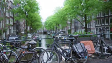 Bisikletler Amsterdam 'da, yemyeşil ağaçların altında, geleneksel Hollandalı evlerle birlikte sakin bir şehir arkaplanı oluşturan pitoresk bir kanal boyunca dizilmişti..