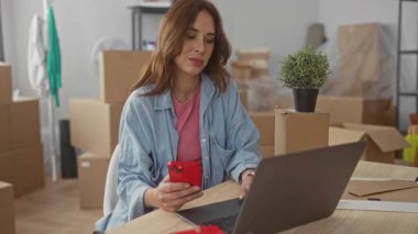 Yeni evinde akıllı telefon ve dizüstü bilgisayar kullanan bir kadın. Arka planda boş kutular var. Bu da yeni oturma odasında rahat ve modern bir oturma odası olduğunu gösteriyor..