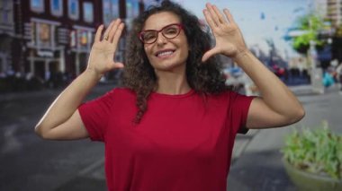 Kırmızı tişörtlü İspanyol genç kadın, kalabalık bir şehir caddesinde avuçlarını açmış binalar ve ekicilerle dolu, neşe dolu..