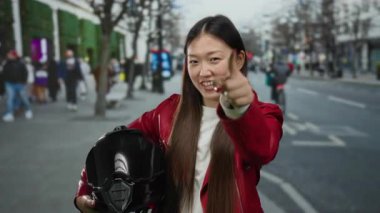 Kırmızı ceketli gülümseyen kadın, kalabalık şehir caddesini işaret ederken miğfer tutuyor..