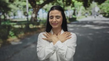 Güneşli bir parkta, yeşil ağaçlarla duygularını ifade eden, beyaz bir gömlek giyen, mutluluk ve tefekkür gösteren, sakin bir açık hava anında yakalanan bir kadın..