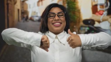 Gözlüklü, gülümseyen genç Latin kadın şehir sokaklarında başparmak kaldırma ve baş parmak kaldırma hareketleri yapıyor, şehir arka planında büyük boy güven gösterisi yapıyor..