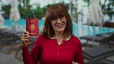 Woman smiling holding mexico passport by a poolside resort, wearing glasses and red shirt, showcasing an outdoor vacation atmosphere, blending travel and relaxation.