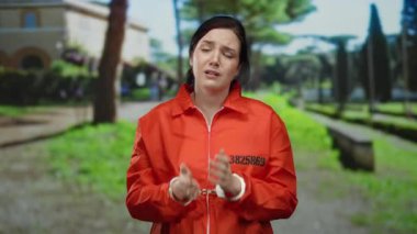 Hapishane tulumlu esmer kadın, kelepçe takıyor, yeşil bir park ortamında açık havada yalvarıyor, duygularını ve savunmasızlığını ifade ediyor..