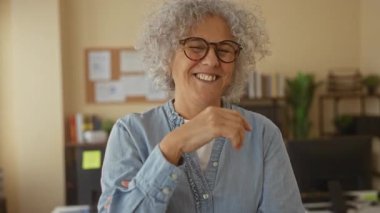 Ofiste gülümseyen gri saçlı ve gözlüklü bir kadın, rahat bir iş ortamında kitaplar ve bitkilerle çevrili..