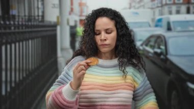 Renkli kazaklı genç kadın, yoğun caddede çerezi incelikle inceliyor. Şehir dışından geçen arabalar, derin düşüncelere dalmış bir ruh halini yansıtıyor..