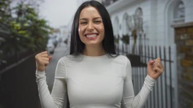 İspanyol kadın şehir sokaklarında sevinçle kutlama yapıyor, açık şehir ortamında yumruklarını sıkarak heyecan gösteriyor, geniş bir gülümsemesi ve beyaz gömleği var..