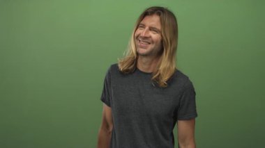 Sarı uzun saçlı, koyu gri tişörtlü yeşil stüdyoda gülen yüzünü gösteren, kaygısız neşe veren bir adam..