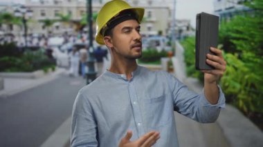 Açık şehir çevresi ve inşaat konseptini gösteren tablet tablet tabletleri kullanarak şehir caddesinde video görüşmesi yapan genç adam..