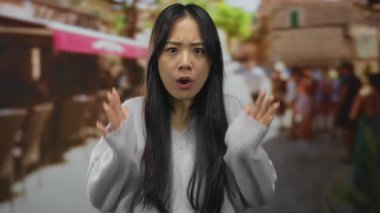 Gri kazaklı genç bir kadın, arka planı bulanık, hareketli çevreyi keşfederken şaşkınlığını ve merakını ifade eden, yoğun bir açık pazar caddesinde duruyor..