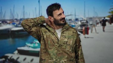 İspanyol üniformalı, sakallı bir adam tekneleri görünen hareketli bir limanda duruyor, parlak mavi gökyüzünün altında boynuna masaj yapıyor gibi görünüyor..