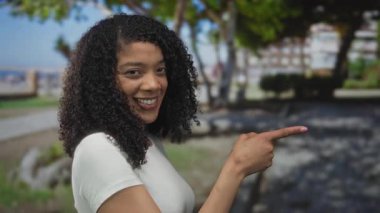 Gülümseyen kadın, şehir parkındaki güneşli nehir geçidini işaret ediyor; merak keşfi bağlantısı.