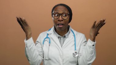 Kadın doktor şaşkınlığını ifade ediyor. Elleri kahverengi arka planda, gözlük ve steteskop takıyor.