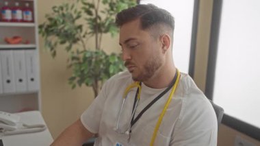 Sakallı ve steteskoplu genç adam hastane ofisinde oturmuş klinik ortamında dizüstü bilgisayarla çalışıyor..