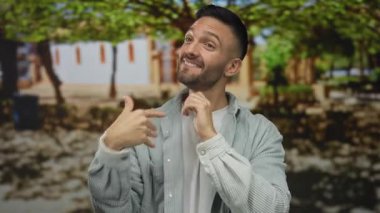 Güneşli bir şehir sokağında ağaçların ve binaların bulanık arka planıyla enerjik bir şekilde konuşan İspanyol genç adam..