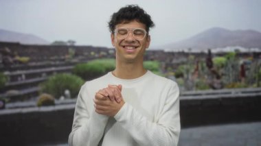 Adam, yıpranmış taş kalıntıları ve bir bina duvarının önündeki berrak bardaklardan ellerini sıkar ve gülümser..