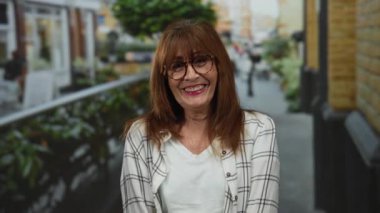 Canlı bir şehir caddesinde gülümseyen bir kadın, mutluluk ve tarz sergiliyor, gözlüklü ve ekose gömlekli bir şehir ortamında doğal ışıklandırma altında son sınıf zarafetini canlandırıyor..