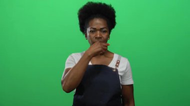 Önlüklü kadın yeşil arka planda sessizliğini koruyor, eli ağzının kenarında sessiz bir mesaj veriyor, iletişimi ve ifadeyi izole bir ortamda vurguluyor..