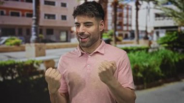 Öğleden sonra güneşli bir şehir sokağında yumruklarını sıkan ve enerjik bir şekilde gülümseyen İspanyol genç adam..