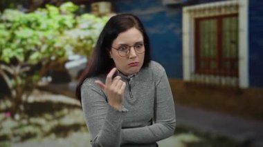 Esmer kadın, açık havada çeşitli duygular ifade ediyor. Gözlük takıyor, yeşil yapraklı, bulanık bir şehir geçmişine karşı meraklı ve düşünceli bir bakış atıyor..