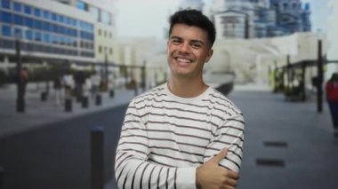 Şehir sokaklarında gülümseyen genç bir adam modern bir arka planda, çizgili bir gömlek giyiyor, mutluluk ve dostluk duygusu taşıyor..