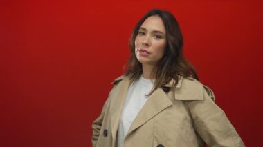 Bej renkli trençkotlu genç İspanyol kadın çarpıcı bir portrede canlı kırmızı bir arka plan, somut bir stil ve modern zarafet karşısında duruyor..