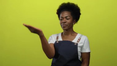 Önlüklü bir kadın avucunu işaret ederek izole edilmiş sarı bir arka plana karşı duruyor, odaklanmış bir ifade ve açık hareketler sergiliyor..