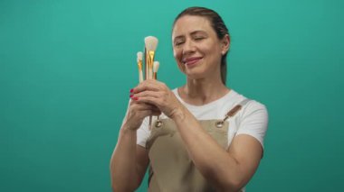 Elinde boya fırçası tutan, bej önlük takan ve duvarları gök mavisi olan bir stüdyoda gülümseyen bir kadın..