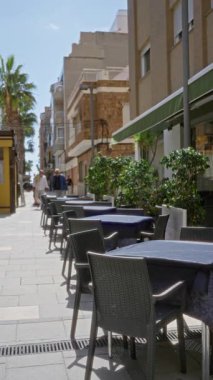 Torrevieja 'da, içi boş masaları ve güneşli gökyüzünün altında sandalyeleri olan, palmiye ağaçları ve konut binalarıyla çevrili bir sokak kafesi manzarası..