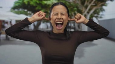 Genç İspanyol kadın dışarıda gözleri kapalı ve kulakları kulaklarında çığlıklar atıyor. Arka planda bulanık yeşil ağaçlarla hayal kırıklığını dile getiriyor..