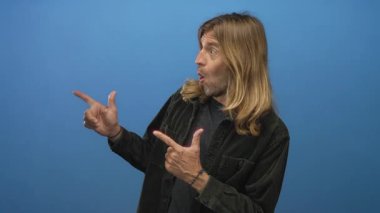 Sarışın, uzun saçlı, işaret parmaklarıyla mavi stüdyoyu işaret eden ağzı açık bir adam..