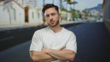 Sakallı genç adam, güneşli bir şehir caddesinde kollarını kavuşturarak, açık hava ortamının güvenini taşıyor..
