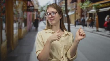 Meşgul bir şehir caddesinde göğsünü gösteren başparmakları gözlüklü ve bej gömlekli genç sarışın kadın, güven gururu pozitifliği güçlendirme.