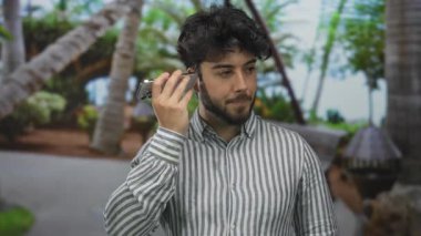 Sakallı genç adam güneşli bir park ortamında sesli mesaj dinler. Çevresi yemyeşil bir bitki örtüsüyle çevrilidir. Rahatlamış ve açık havada bir konuşmayla meşguldür..