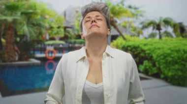 Beyaz gömlekli, gözleri kapalı ve başı eğik bir kadın binanın yanında mavi havuz kenarında duruyor..