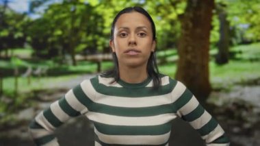 Güneşli park ortamında sinirli bir kadın çizgili kazağıyla canlı yeşil bir arka plana karşı duygularını ifade ediyor..