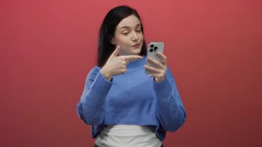 Akıllı telefon tutan kadın kırmızı arka plana başparmak işareti veriyor mavi süveter giyiyor ve kendine güvenen ifade dijital ortamda pozitifliği ve bağlantıyı sembolize ediyor