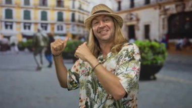 Uzun saçlı, saman şapkalı ve tropikal tişörtlü sarışın bir adam şehir meydanını bulanık yayaların arasından işaret ediyor..