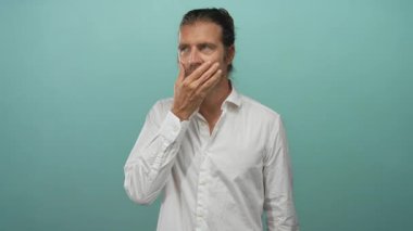 Beyaz gömlekli adam stüdyoda yanağına turkuaz arka planla dokunuyor. Düşünceli bir hareket sergiliyor..