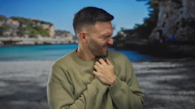 Arka planda bulanık bir deniz kıyısı ve deniz kayalıklarıyla plajda göğsünü sıkıntı içinde tutan İspanyol genç bir adam..