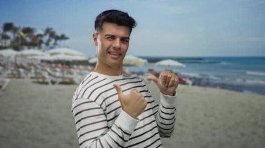 Genç adam kumsalda gülümsüyor, çizgili gömlek giyiyor, arka planda deniz ve şemsiyeler var, baş parmaklarını kaldırıyor, rahat bir sahil havası yakalıyor..