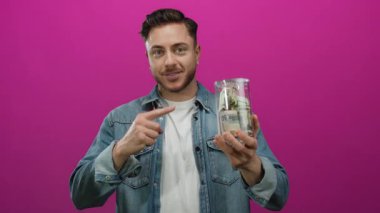 Sakallı genç bir adam, izole edilmiş pembe bir arkaplanı işaret eden bir kavanoz dolusu dolarımızı tutuyor. Kot ceket giyiyor ve kameraya içten bir şekilde gülümsüyor.