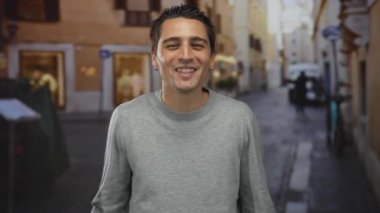 Gündelik kıyafetli genç İspanyol bir adam, güneşli bir günde, şehir manzarasıyla çevrili neşeli bir titreşim yayarak, bir şehir caddesinde şakacı bir şekilde gülümsüyor..