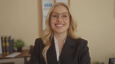 Ofisteki kadın kendine güvenen gözlüklü ve takım elbiseli sarı saçlı, kitaplı ve arka planda bitki yetiştiren modern ve profesyonel bir atmosfer yaratıyor..