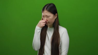 Burnunu çimdikleyen bir kadın canlı yeşil arka planda yakın planda uzun saçlı ve beyaz süveterli kadın duygularını dışa vuruyor..