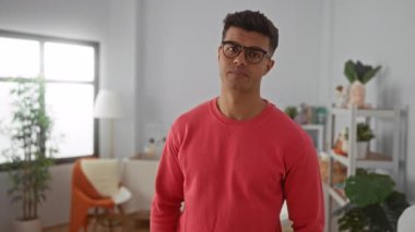 Kırmızı kazaklı ve gözlüklü genç adam kapalı bir odada rahat dekor ve bitkilerin yer aldığı çapraz kollarıyla güvenle gülümsüyor..