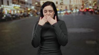 Şehir ışıkları ve bulanık arka planla kalabalık caddede endişeli olduğunu ifade eden esmer kadın canlı bir şehir ortamında duygusal bir tepkiyi işaret ediyor..