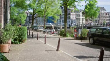 Amsterdam 'daki bulanık sokak manzarası ağaçlar, kanallar ve Hollanda' daki kaldırım taşı patikası boyunca park edilmiş bisikletlerle güneşli bir şehir manzarası gösteriyor..