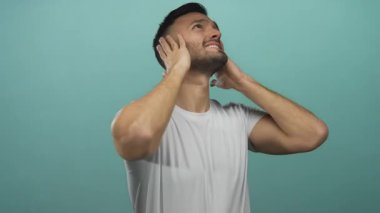 Genç adam, mavi bir arka plana karşı hayal kırıklığına uğramış bir ifadeyle kulaklarını kapatıyor, beyaz bir gömlek giyiyor ve stresli ve endişeli görünüyor..