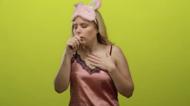 Pembe gecelikli genç sarışın kadın. Tavşan uyku maskesi takmış. Parlak sarı bir arka plana doğru öksürüyor..